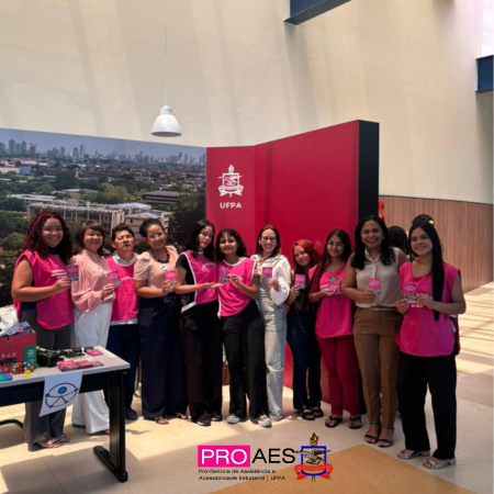 Grupo de mulheres posa sorrindo em um espaço interno da UFPA, a maioria vestindo rosa e segurando materiais informativos. Ao fundo, há um painel com a sigla da universidade. Na parte inferior, aparece a identidade visual do PROAES, relacionado à assistência estudantil.